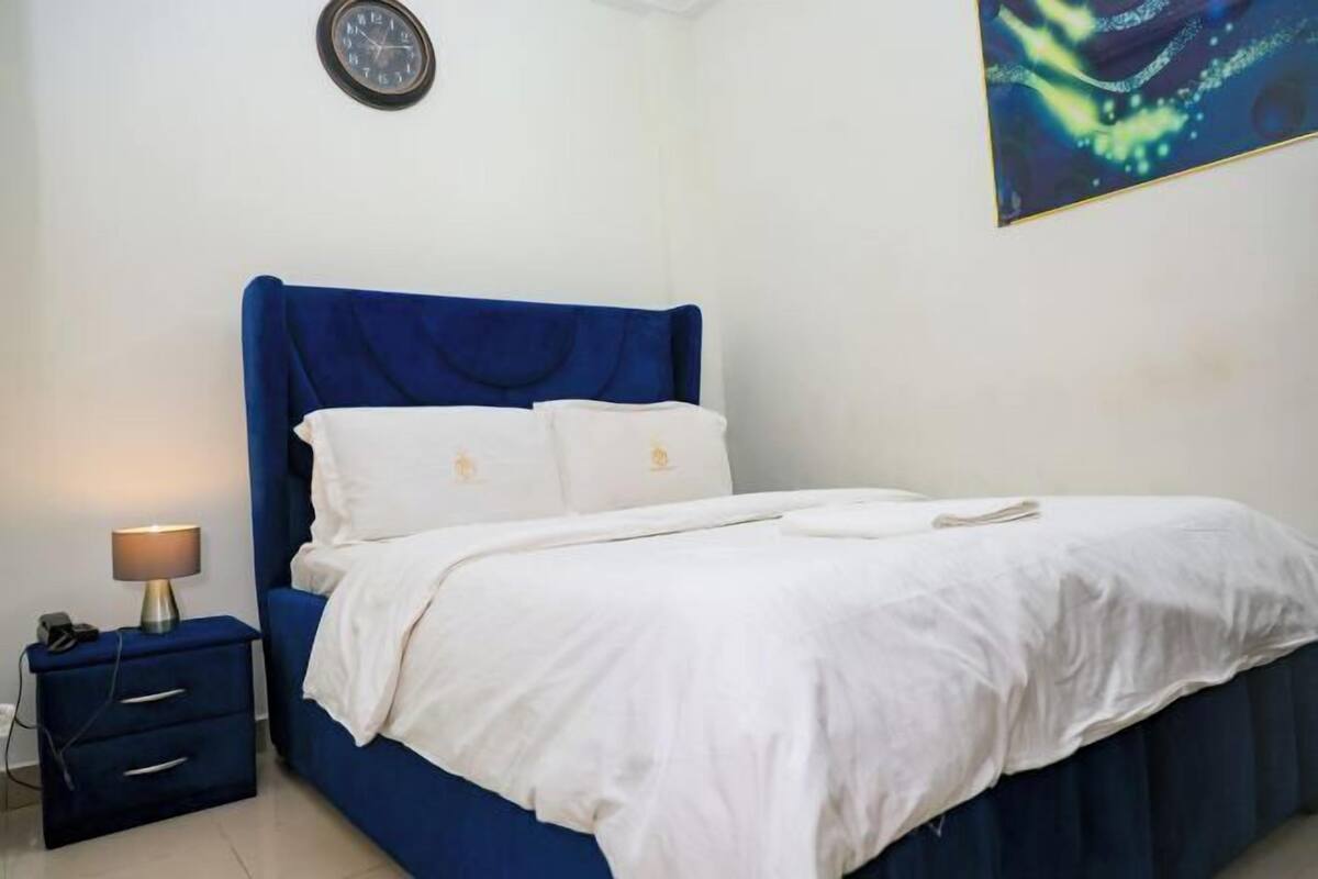 Une chambre à coucher avec une tête de lit bleue, du linge de lit blanc, une lampe de chevet et une horloge au mur.