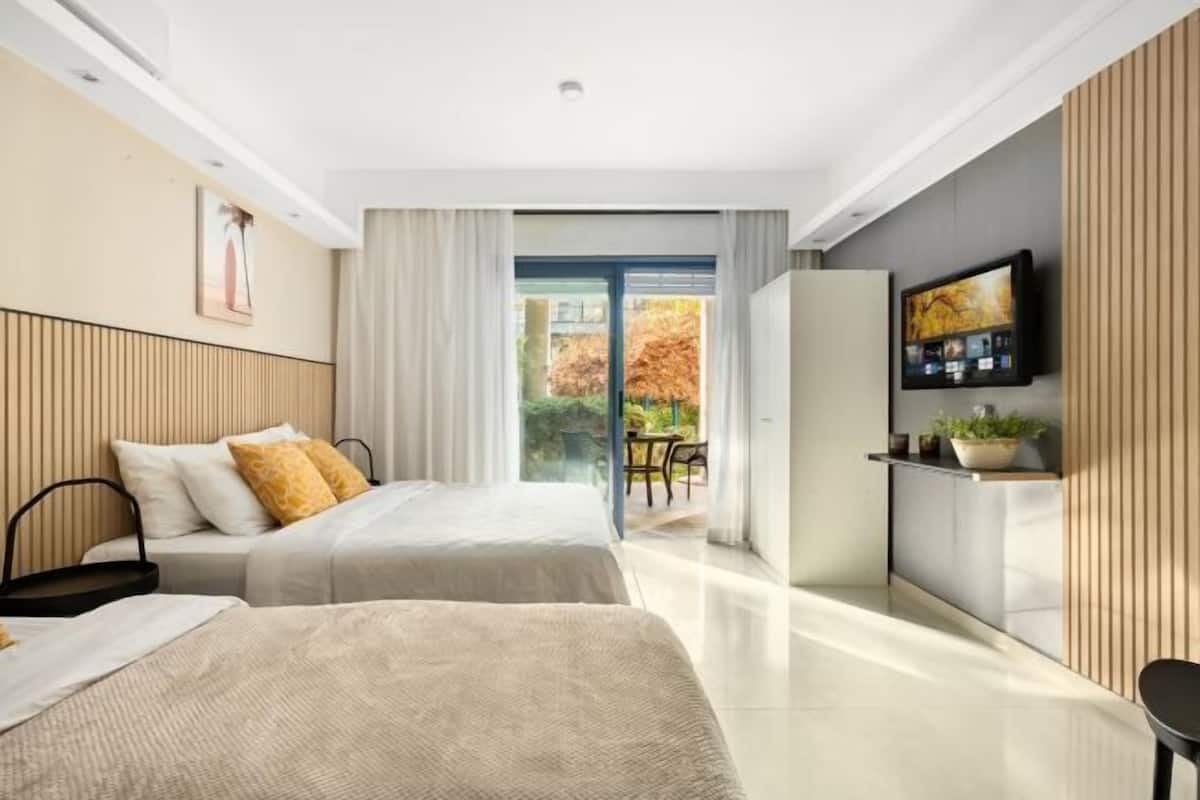 A modern hotel room with two beds, a TV, a small kitchen area, and a sliding glass door leading to an outdoor seating area.