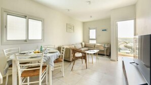 A spacious living area with a dining table, chairs, a sofa, and a TV. - Skyline 1BR Apartment with City and Sea View (Herzliya)
