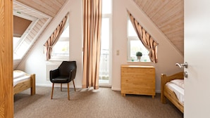 A bedroom with a sloped ceiling, a wooden dresser, a black chair, a bed, and a window with curtains.