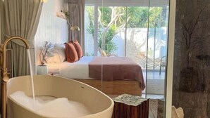 A modern bedroom with a large bathtub, a bed with a brown headboard, and a view of greenery outside. - Wynn resort (Bang Lamung)