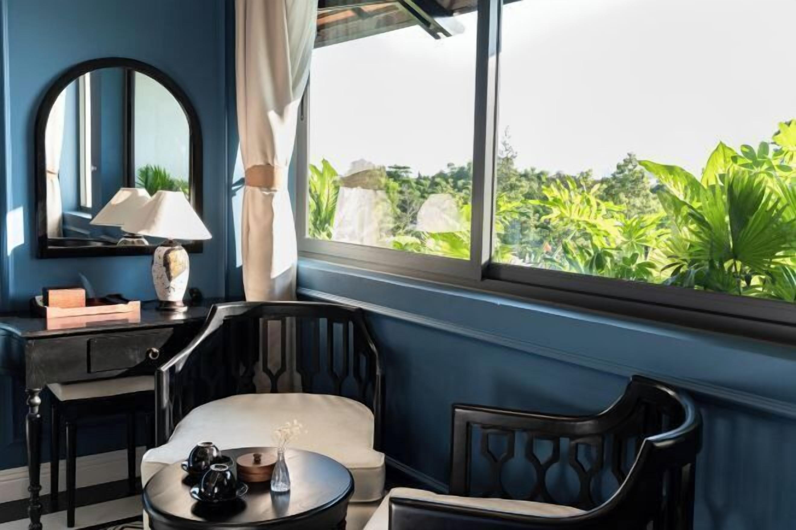 A room with a large window, a mirror, a table with a lamp, and a chair.
