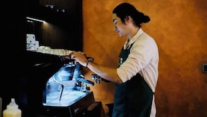 Eine Person bedient eine Kaffeemaschine in einer Küche mit Holzwand und Fenster.