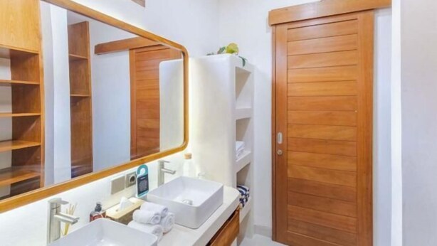 Un baño con un espejo grande, dos lavamanos, armarios de madera y una puerta de madera.