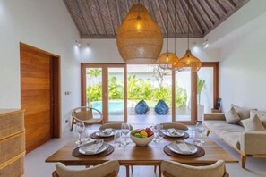 Un comedor con una mesa preparada para cuatro personas, una lámpara de techo de mimbre y vistas a una piscina y follaje.