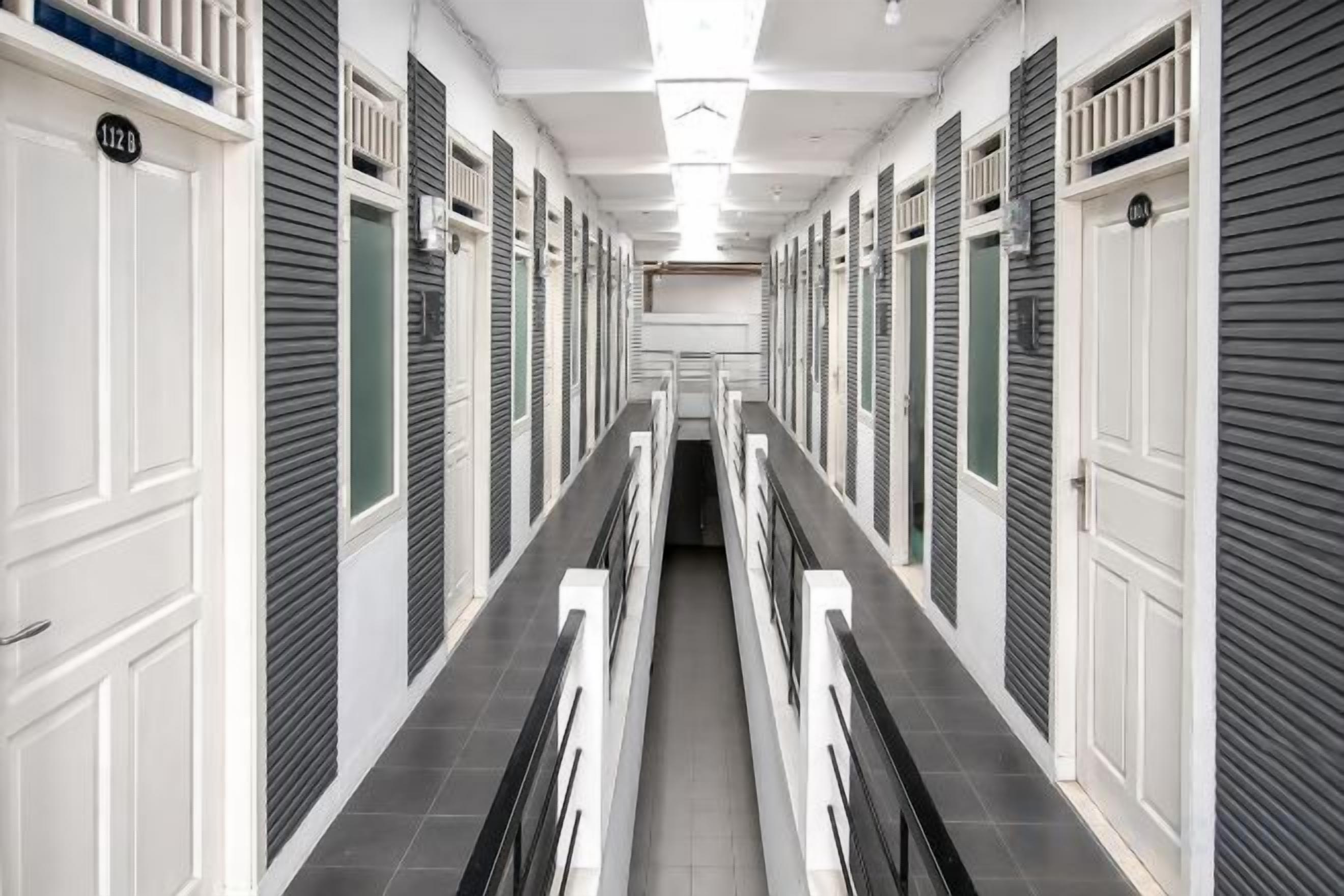 A hotel corridor with closed room doors, a central walkway, and a ceiling light fixture.