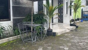Uma área externa com mesas e cadeiras, rodeada por plantas em vasos e piso de pedra.