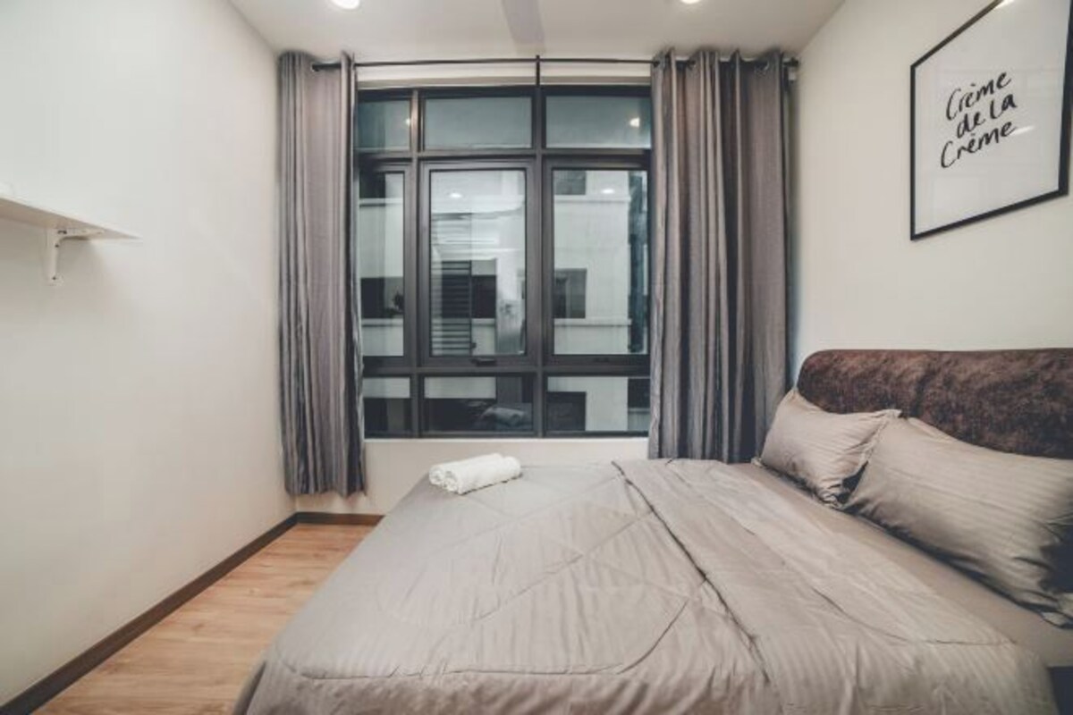 Een slaapkamer met een groot bed, een raam met gordijnen, een citaat in een lijst op de muur en een houten vloer.