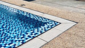 Ein Schwimmbecken mit blauem Mosaikfliesenmuster, umgeben von hellen Fliesen und einer Holzterrasse.