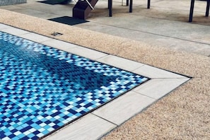 Une piscine dont le fond est recouvert de mosaïques bleues, entourée de carreaux de couleur claire et d’une terrasse en bois.