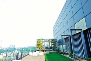 Un bâtiment moderne doté d’une façade en verre, d’un jardin sur le toit avec des chaises blanches et d’une vue sur le paysage urbain.