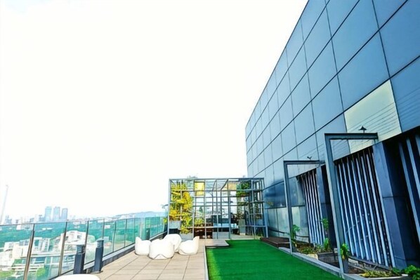 Un edificio moderno con fachada de vidrio, una terraza en la azotea con césped artificial y asientos, y vista panorámica de la ciudad.