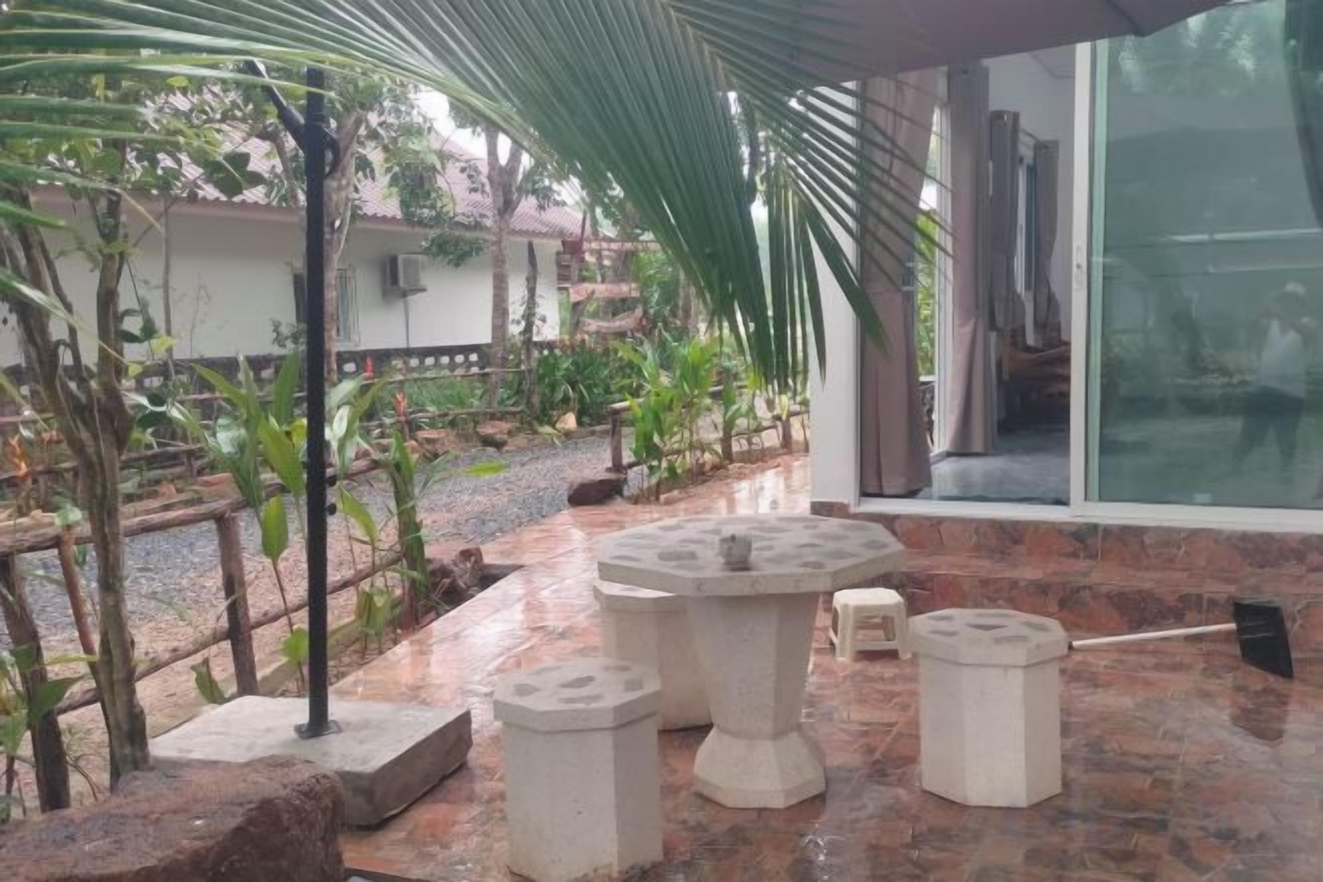 An outdoor patio area with a round table and stools, surrounded by plants and a gravel path.