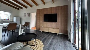 A modern living room with a large TV, wooden panel wall, and a dining area with chairs and a table. - Roseneath Holiday Park (Meerlieu)