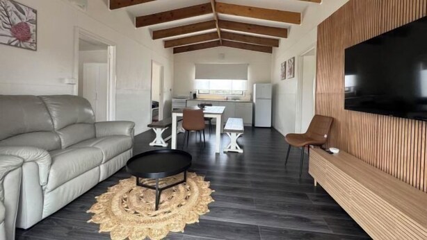 A modern living room with a sofa, a dining area with a table and chairs, a kitchen in the background, and a TV mounted on a wooden wall. - Roseneath Holiday Park (Meerlieu)