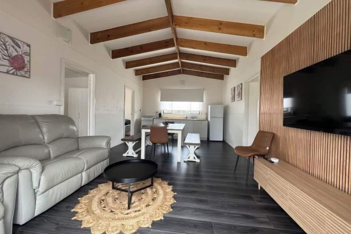 Una sala de estar moderna con un sofá, un comedor con mesa y sillas, una cocina al fondo y un televisor montado en una pared de madera.
