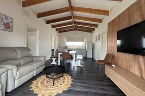 Una sala de estar moderna con un sofá, un comedor con mesa y sillas, una cocina al fondo y un televisor montado en una pared de madera.