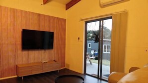 A living room with a flat-screen TV mounted on a wooden panel wall, a sofa, a coffee table, and a sliding glass door leading to an outdoor area. - Roseneath Holiday Park (Meerlieu)