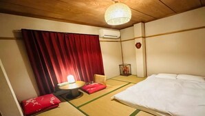 Una habitación con suelo de tatami, una cama, una pequeña mesa con un cojín rojo, un aire acondicionado de pared y una lámpara colgante.