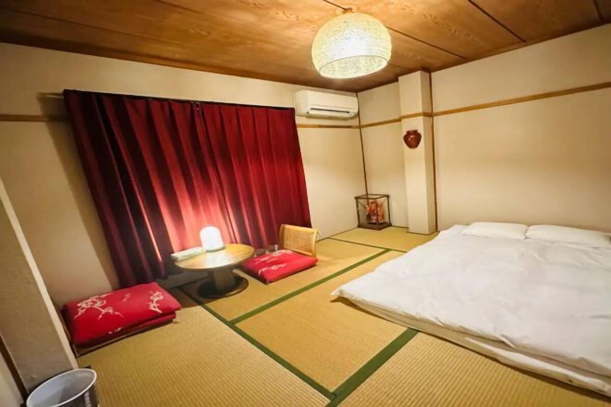 Een kamer met tatami vloerbedekking, een bed, een klein tafeltje met een rode kussen, een wandairconditioner en een hanglamp.