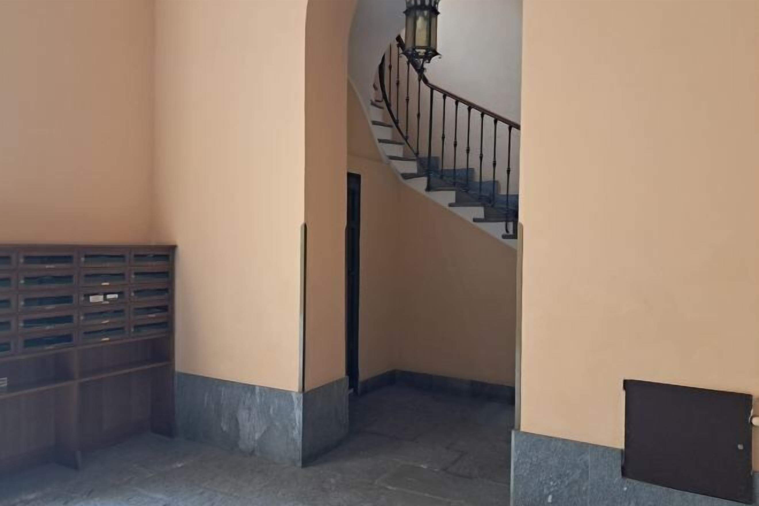 Un pasillo con una escalera curva, barandilla metálica y una lámpara fijada a la pared.