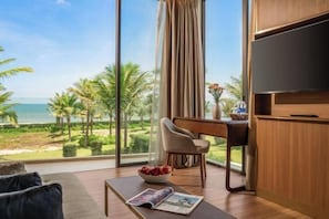 A modern room with a large window overlooking a beach and palm trees. There is a sofa, a dining table with chairs, and a flat-screen TV mounted on the wall. - Legasea Villa Hoi An Private Pool 3Night pick - up (Dien Ban)