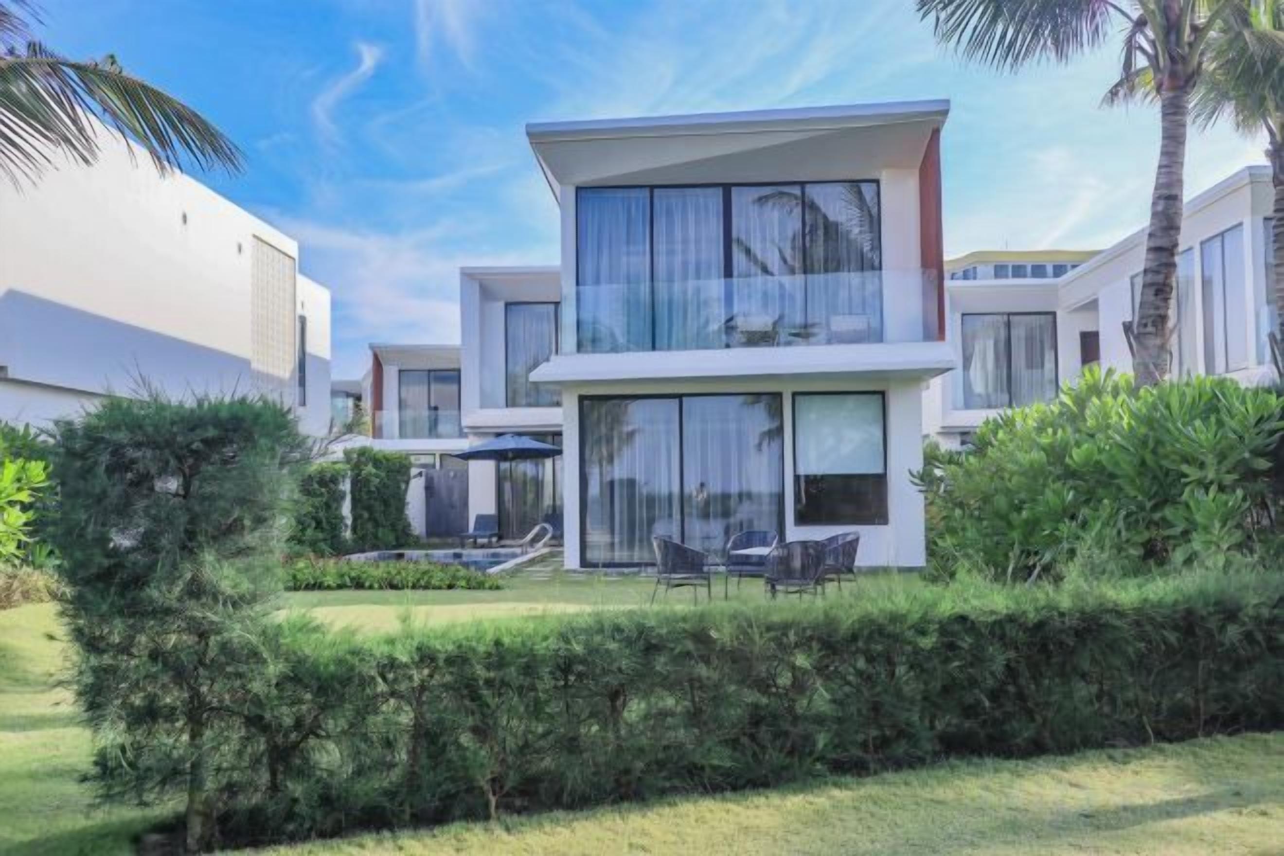 A modern two-story house with a balcony, surrounded by greenery and a clear sky.