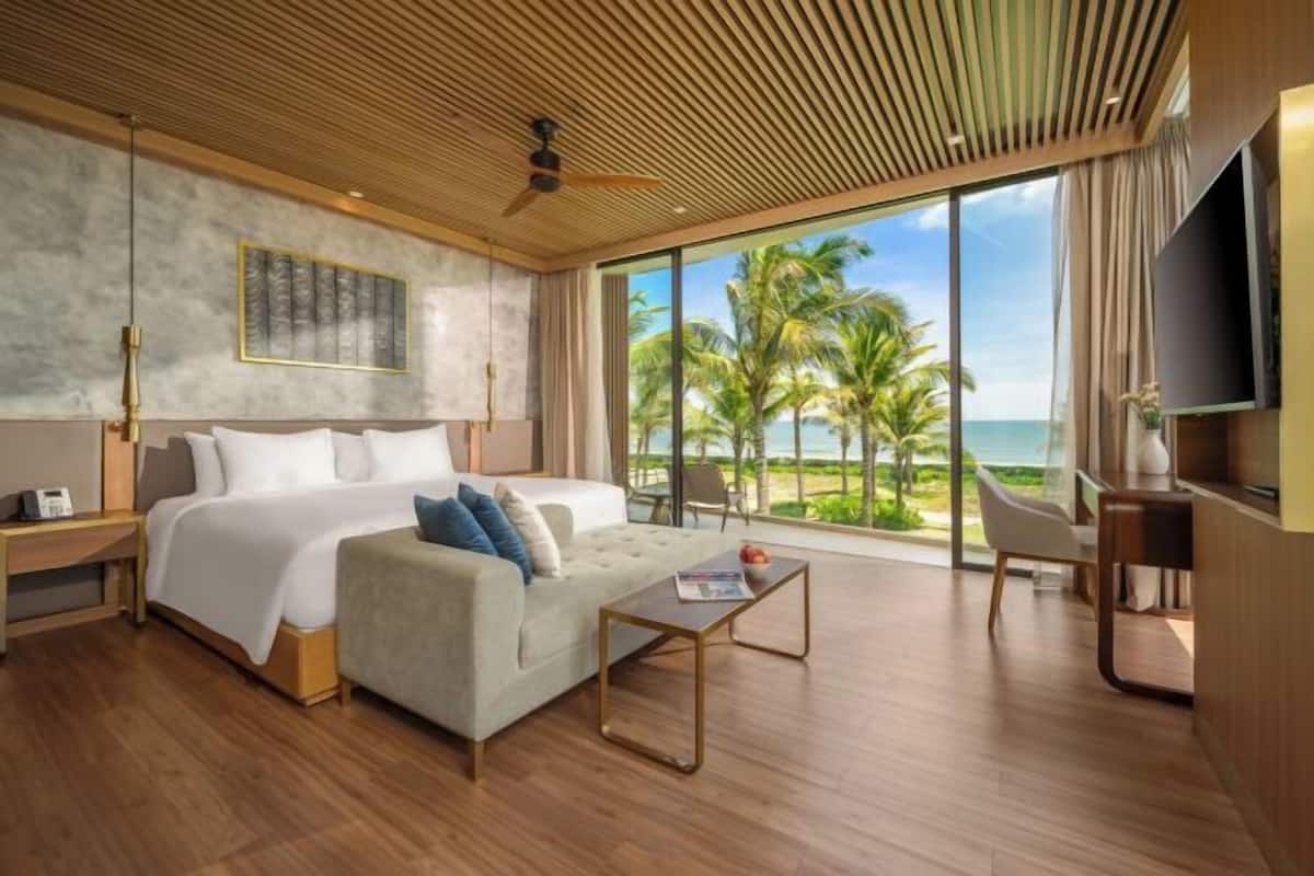 A modern hotel room with a large bed, a sofa, a coffee table, and a TV. The room has a large window with a view of palm trees and the ocean.