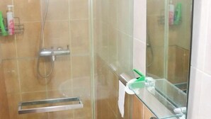 A glass-enclosed shower area with a handheld showerhead, a glass shelf with toiletries, and a glass door. - Taman Sari Sudirman Studio Deluxe B / Near MRT / Central Jakarta (South Jakarta)