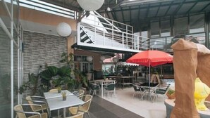 Un área de café al aire libre con mesas, sillas, un paraguas rojo y plantas en macetas.