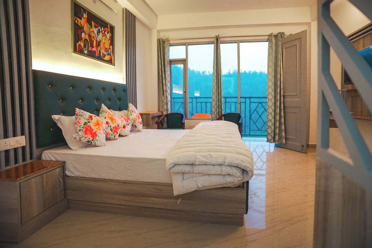 A hotel room with a large bed, a nightstand, a chair, a balcony with a view, and a painting on the wall.