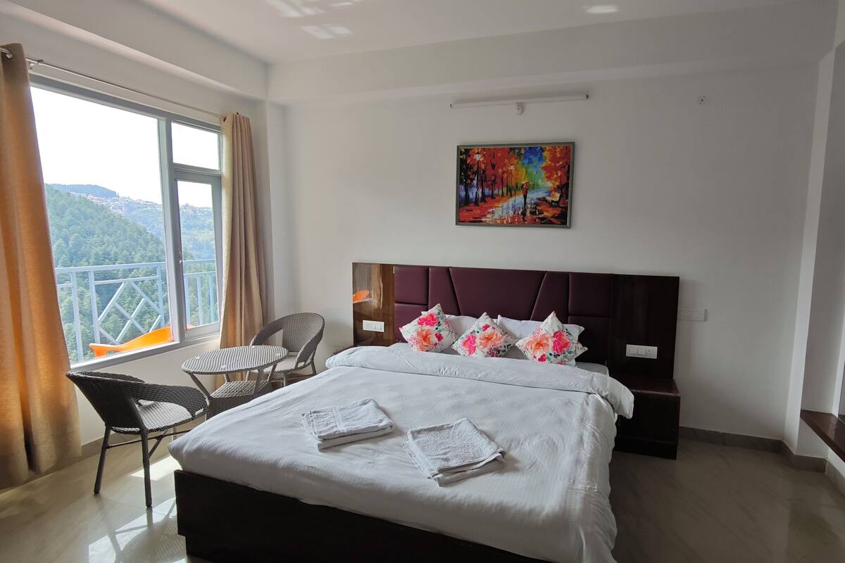 A hotel room with a bed, a chair, a small table, a painting on the wall, and a balcony with a view of the hills.