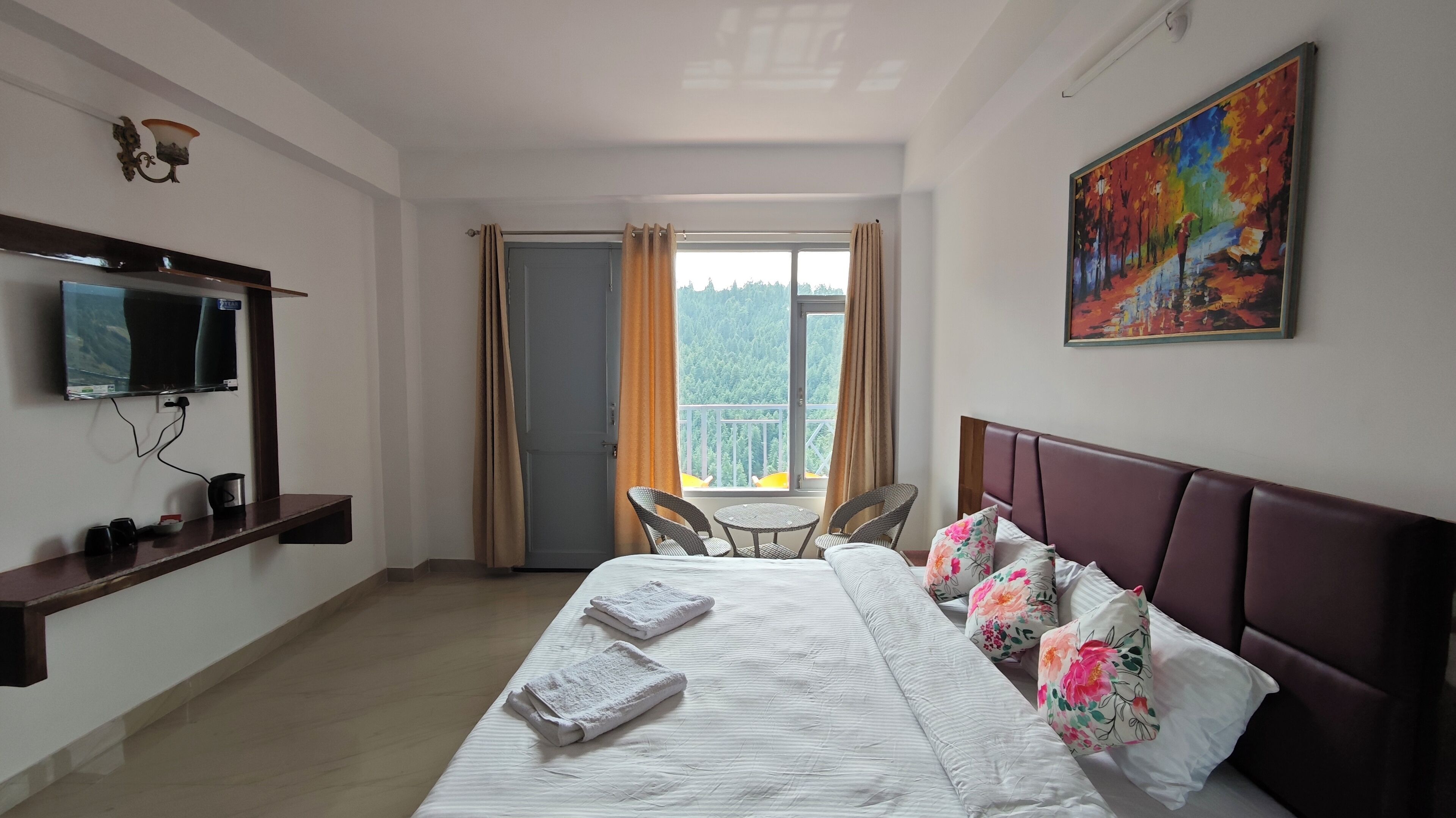 Habitación de hotel con cama, televisor, balcón con mesa y sillas, y un cuadro en la pared.