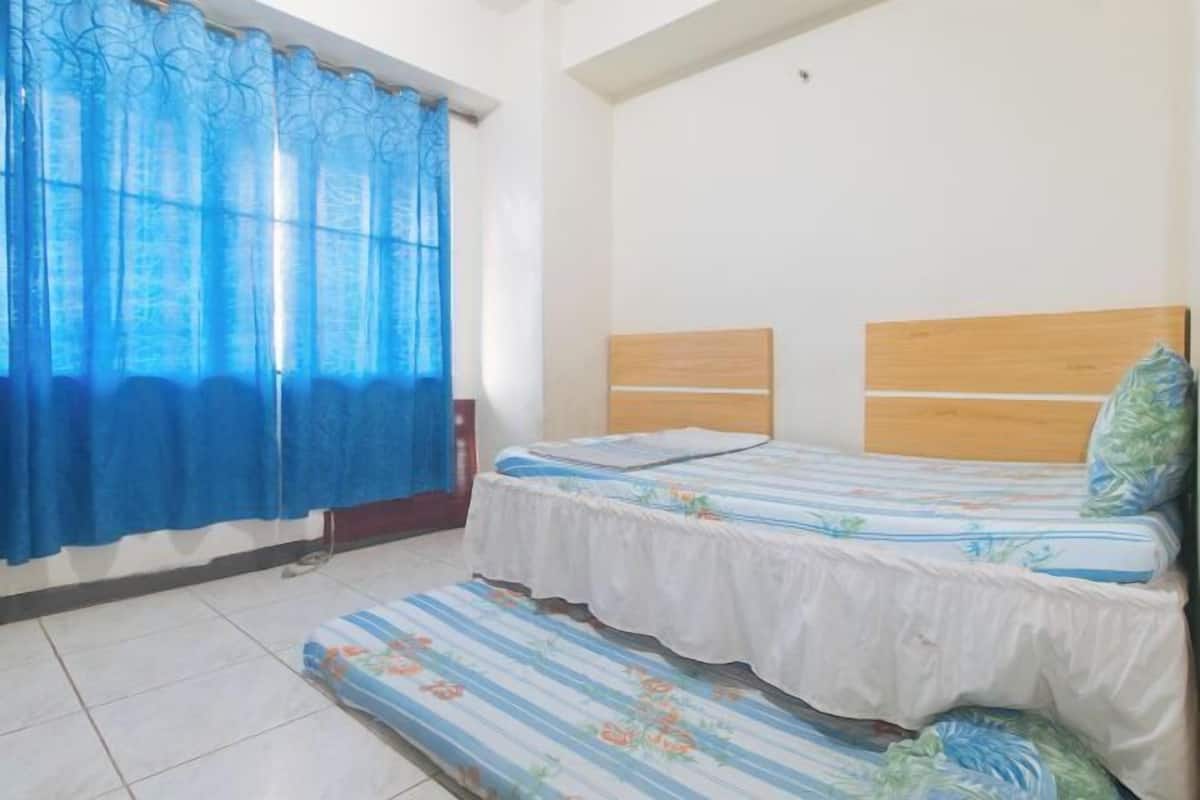 A room with two beds, a window with blue curtains, and a tiled floor.