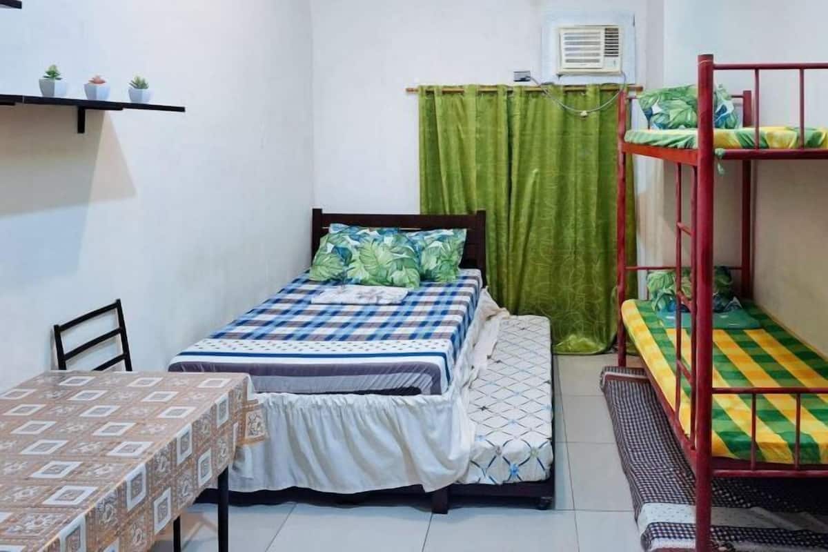 A dormitory room with bunk beds, a single bed, a table with a patterned tablecloth, a green curtain, and an air conditioning unit.