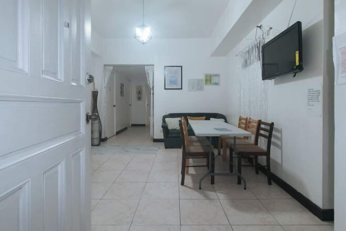 A dining area with a table and chairs, a television mounted on the wall, and a door leading to another room.