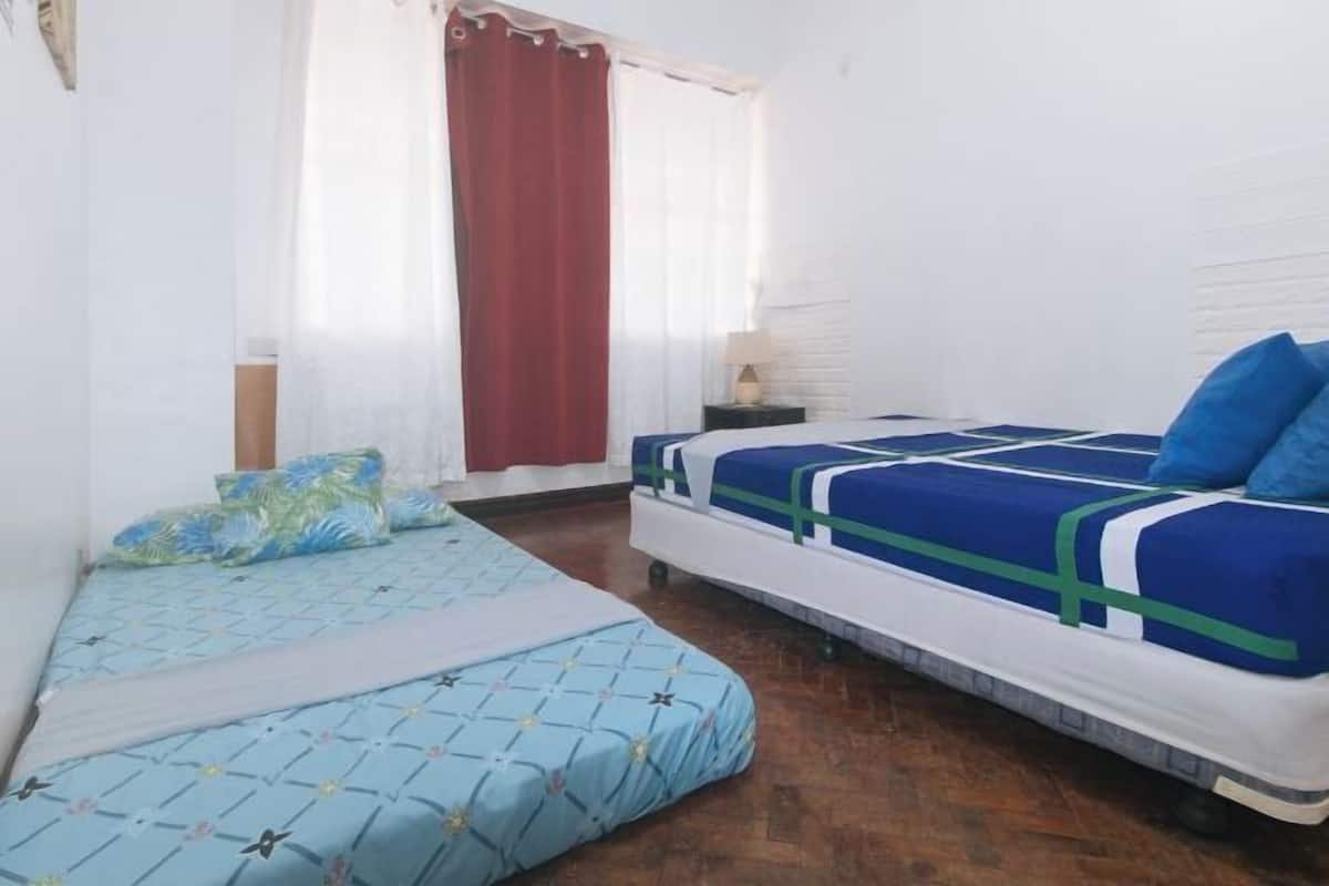 A bedroom with two beds, a wooden floor, a window with curtains, and a bedside table with a lamp.