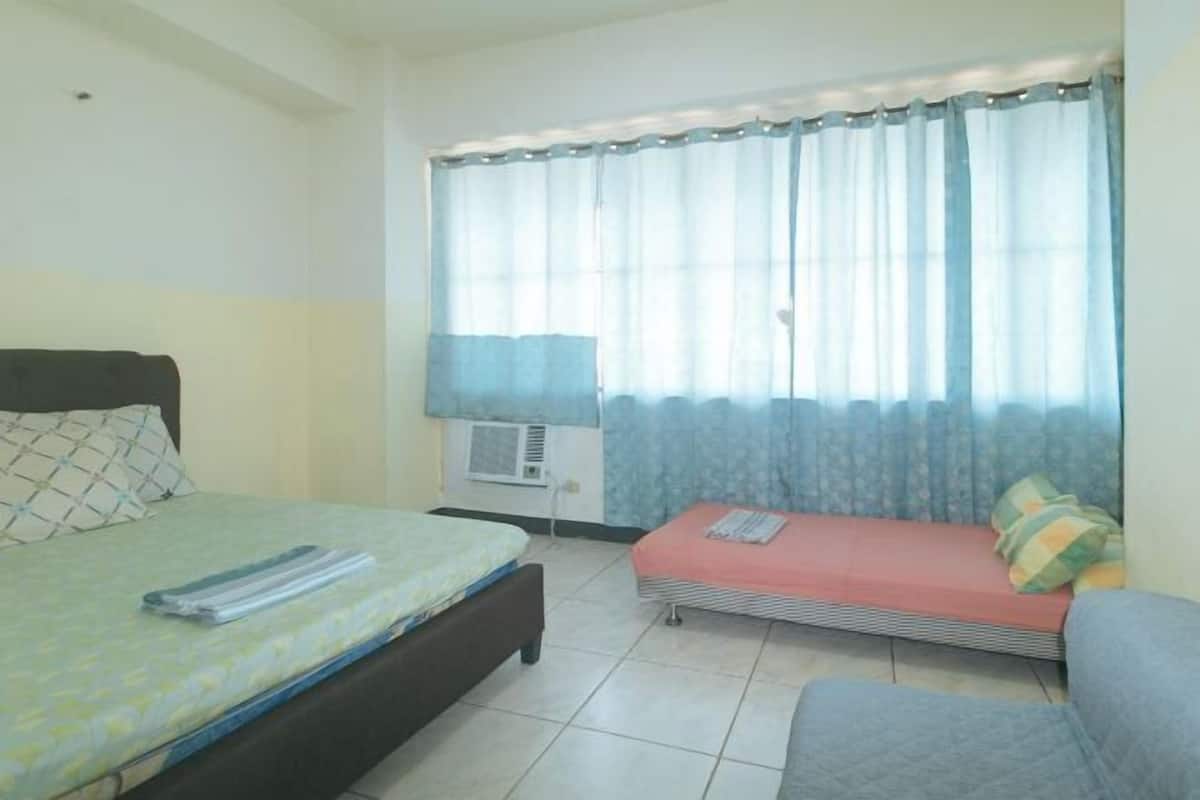 A bedroom with a bed, a sofa, a window with curtains, an air conditioning unit, and a wall-mounted lamp.