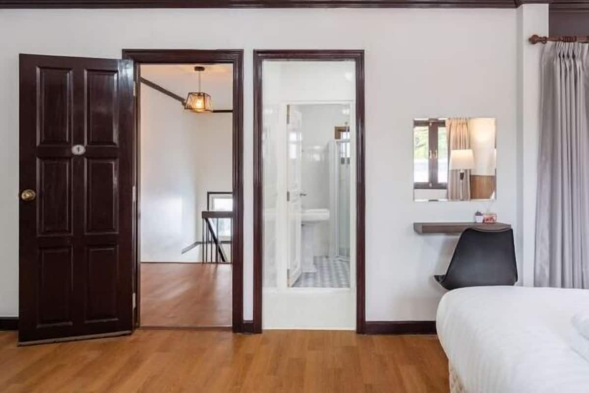 A hotel room with a wooden floor, a bed, a desk with a chair, a mirror, and a window with curtains.