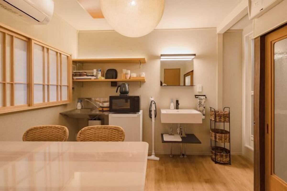 A compact kitchen with a microwave, sink, and small refrigerator. There is a dining table with chairs and a hanging light fixture.