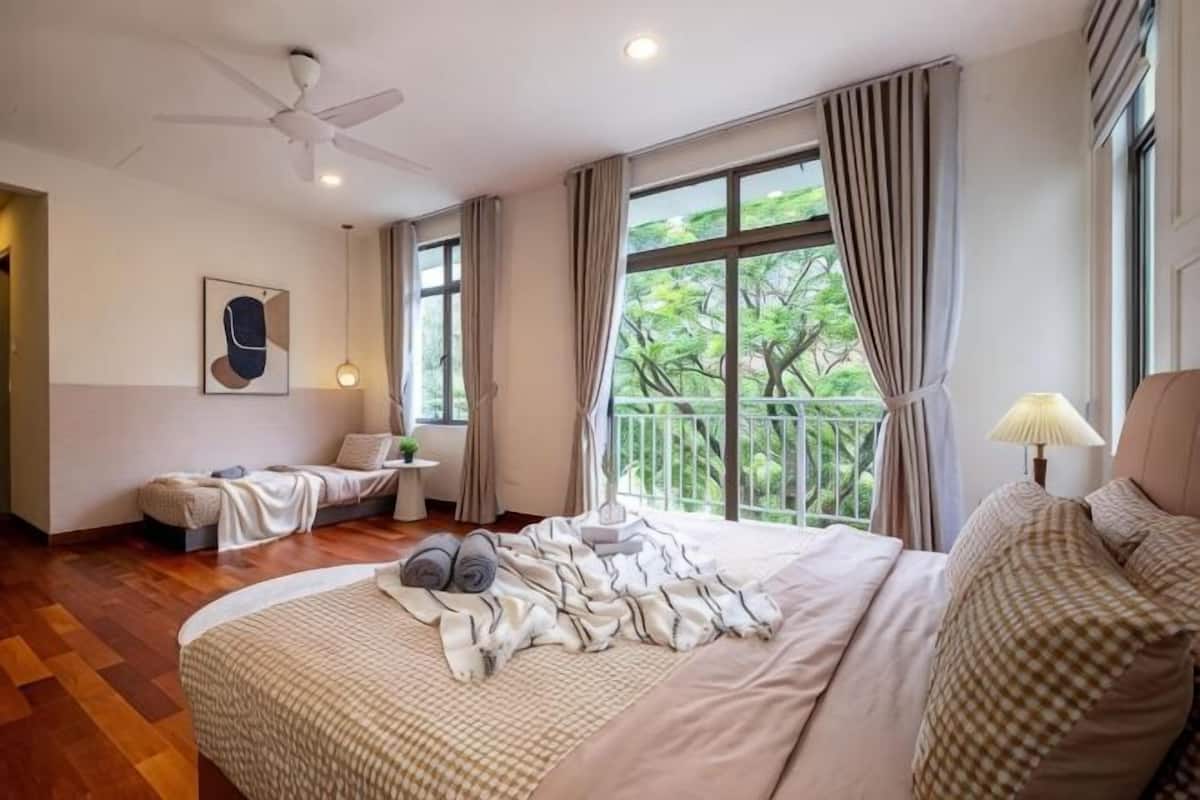 A bedroom with a large bed, a ceiling fan, a window with curtains, a bedside table with a lamp, and a view of greenery outside.