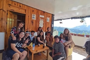 A group of people seated on a wooden bench and chairs outside a building with wooden walls and a view of mountains.