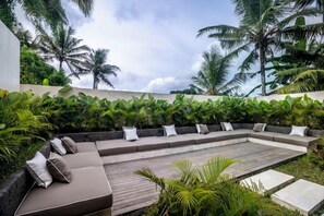 Un espacio al aire libre moderno con terraza de madera, asientos acolchados y exuberante vegetación tropical.