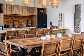 A dining area with a long wooden table and wicker chairs, a kitchen with a wooden backsplash, and decorative wall art. - Villa Zen Paradise by BaliSuperHost (Bali)