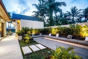 Een modern buitenverblijf met een houten terras, omringd door weelderige tropische planten en palmbomen.