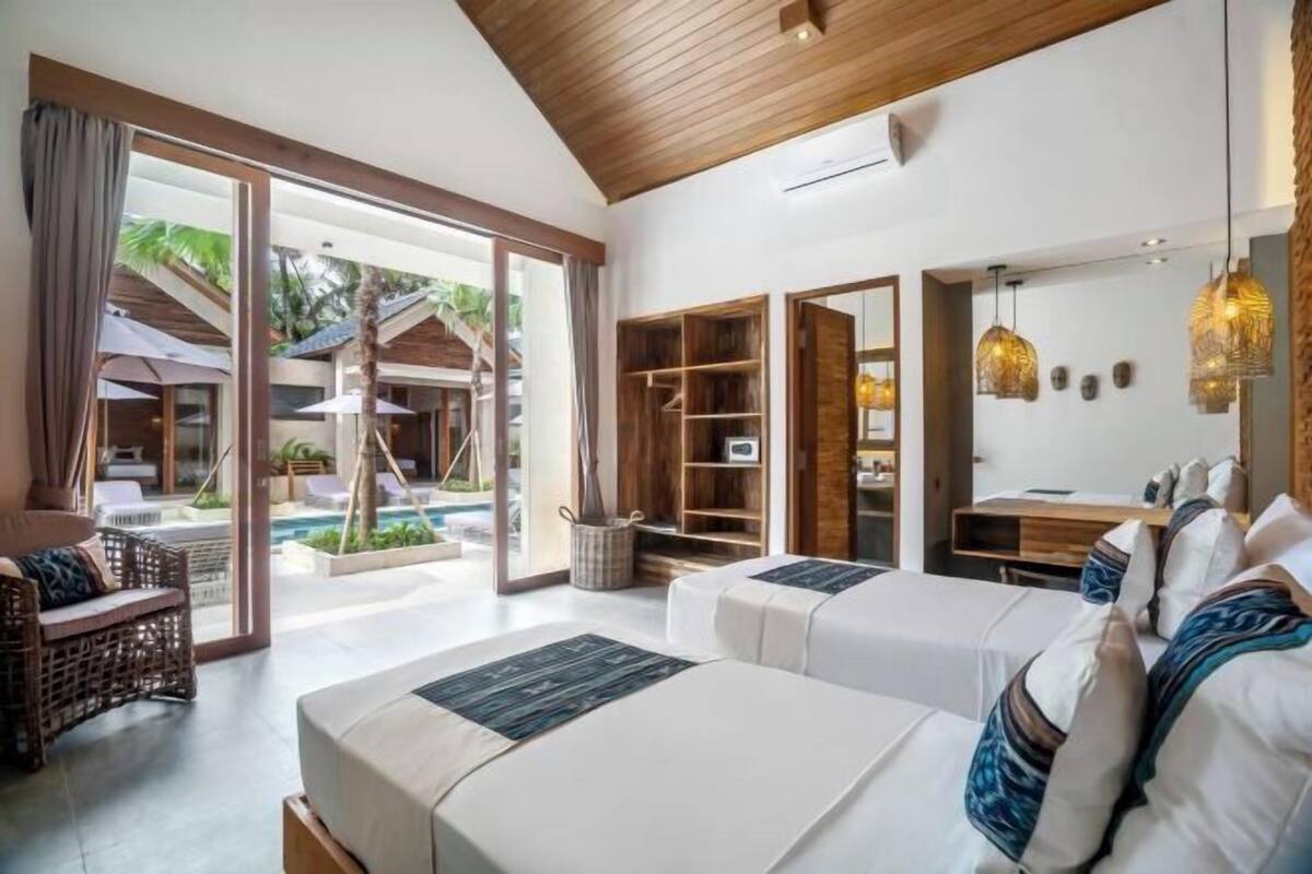 Habitación de hotel con dos camas, una zona de estar con una silla, techo de madera y vista al área de la piscina.