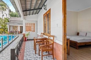 A room with a bed, a small table, and a chair, a wooden door, and a view of a pool.