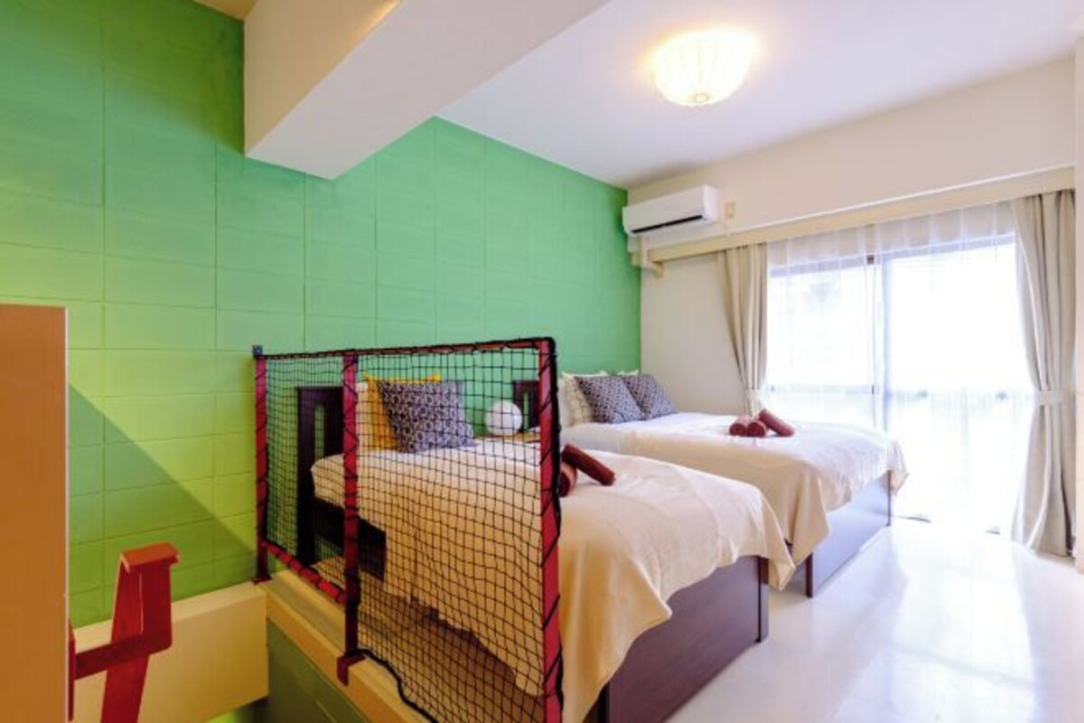 A hotel room with two beds, a green wall, an air conditioning unit, and a window with curtains.