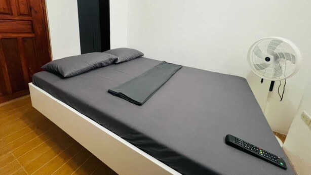 A single bed with a grey mattress and pillows, a remote control on the bed, and a standing fan to the right. - T&A Building  (Pasay)