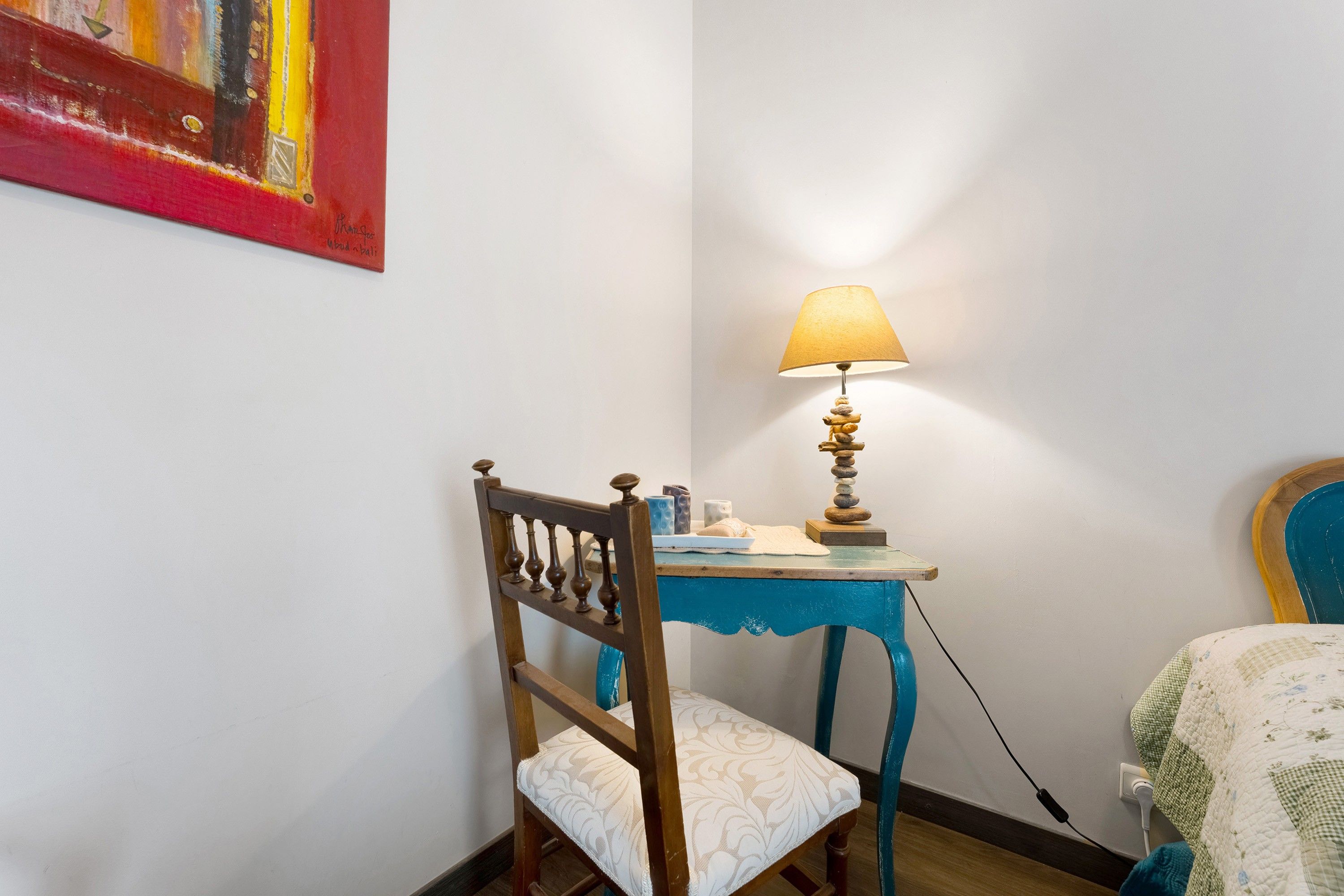 A corner room with a wooden chair, a small table with a lamp, and a painting on the wall.
